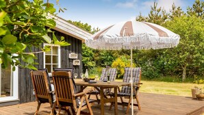 Une terrasse en bois avec une table et des chaises, un parasol et un petit abri de jardin, le tout aménagé dans un cadre verdoyant.