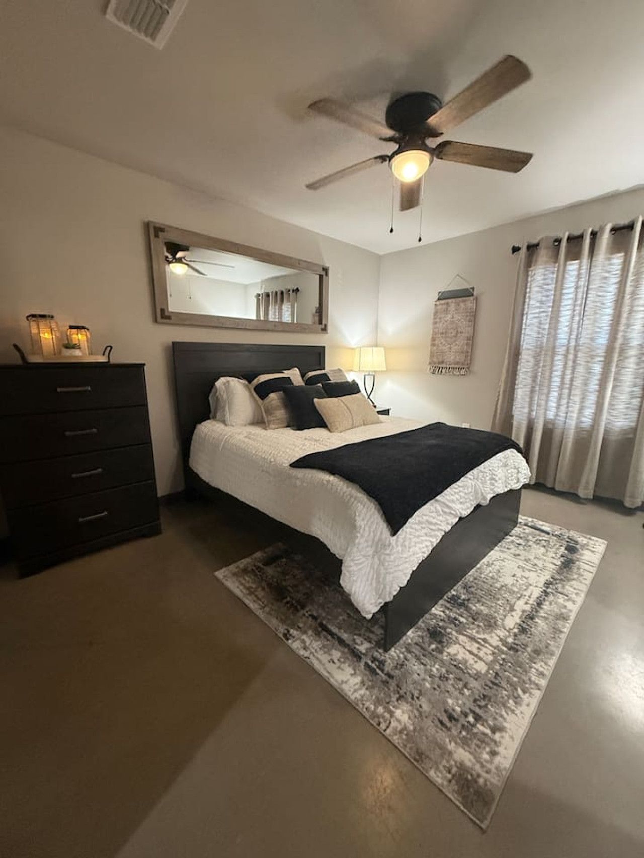 A bedroom with a bed, a ceiling fan, a dresser, a mirror, and a window with curtains.