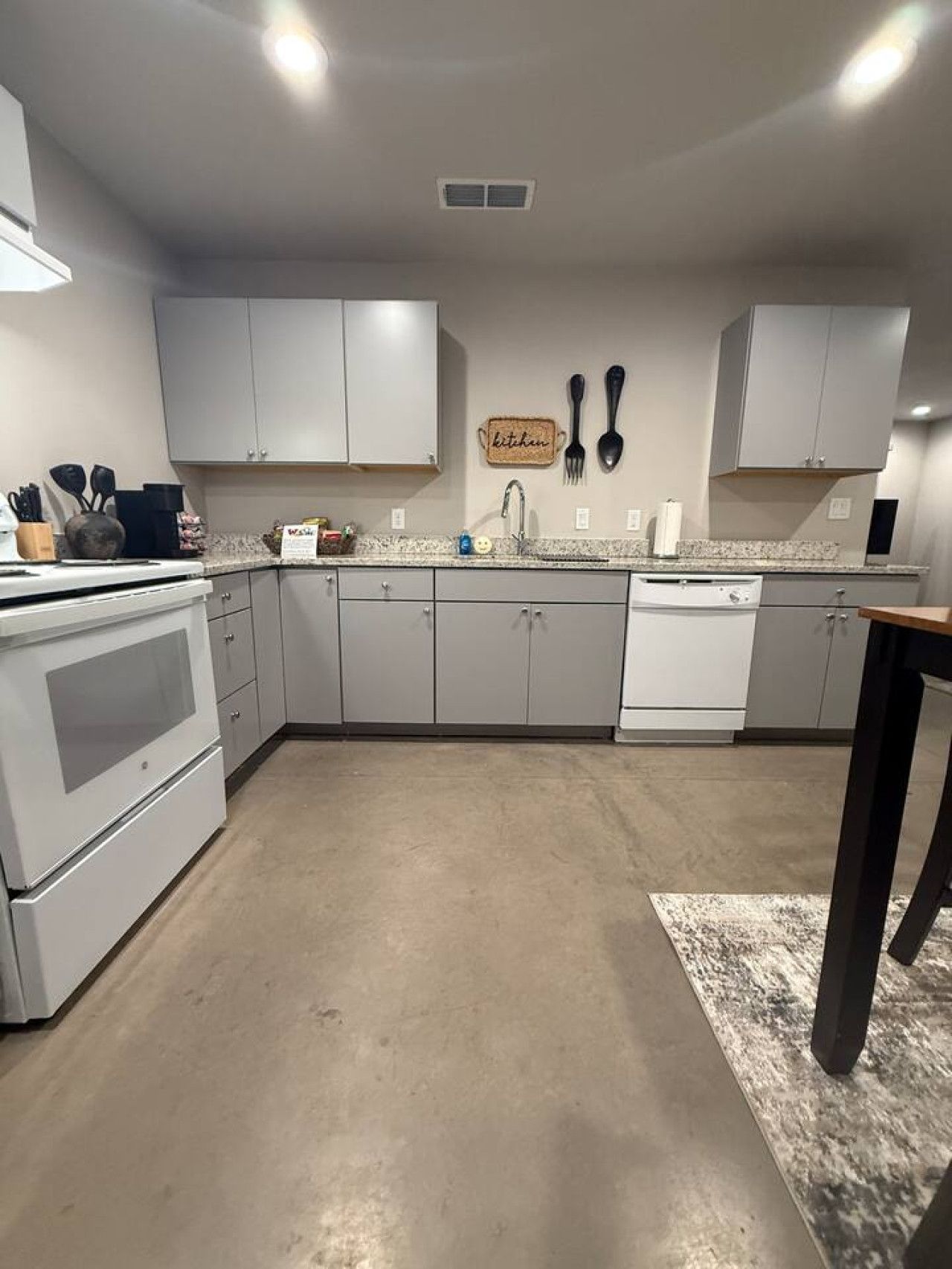 A kitchen with grey cabinets, a stove, a dishwasher, and a sink. There is a kitchen sign on the wall and a rug on the floor.