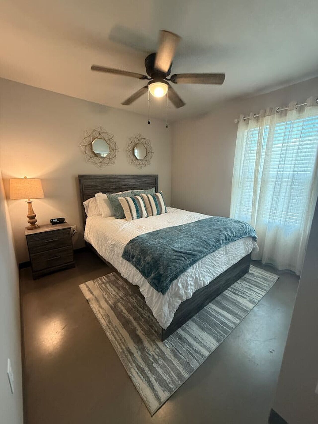 Un dormitorio con cama, mesita de noche, ventilador de techo y ventana con cortinas.