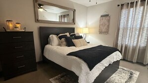A bedroom with a bed, a ceiling fan, a dresser, a mirror, and a window with curtains. - Calming Comfort Stay (Clovis)