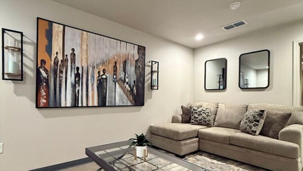 A living room with a large abstract painting, a beige sofa, a glass coffee table, and three wall-mounted mirrors. - Calming Comfort Stay (Clovis)