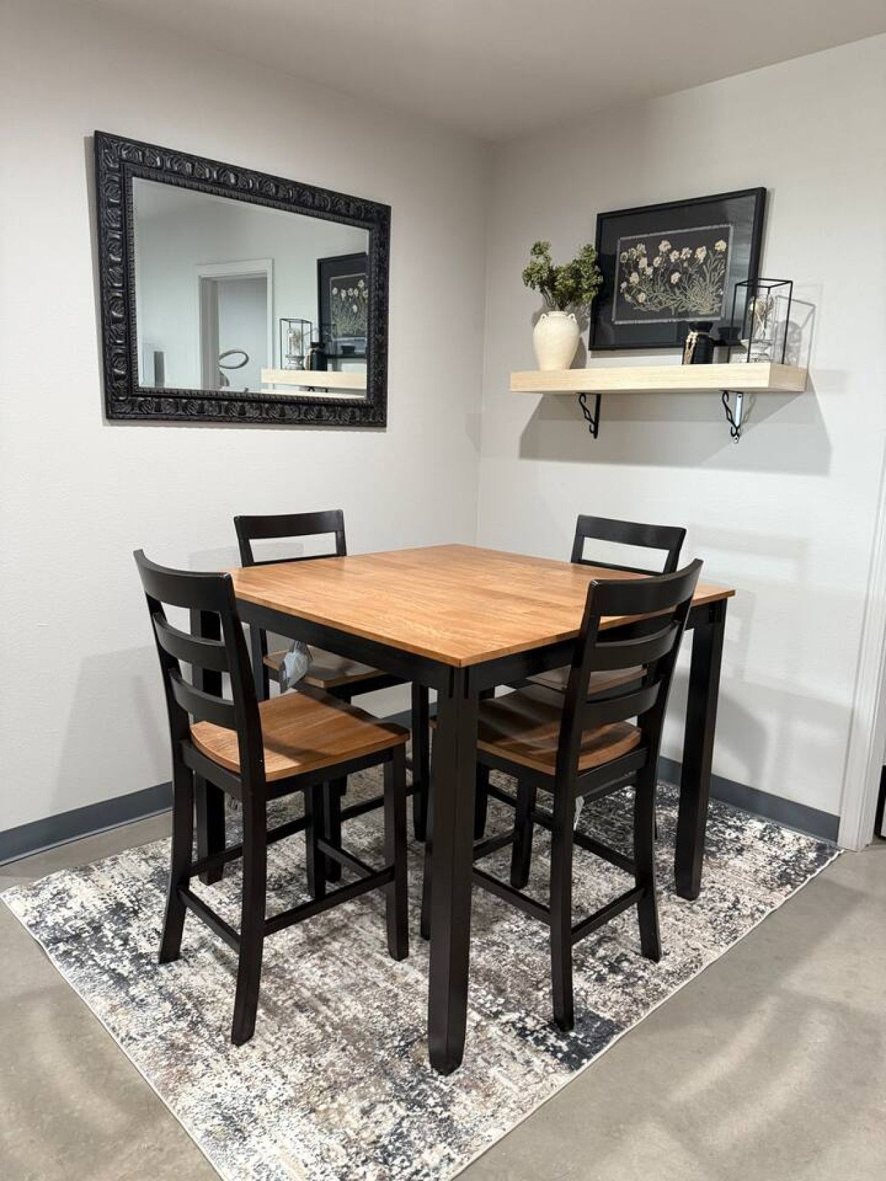 Un comedor con mesa de madera y cuatro sillas, un espejo con marco negro, y una estantería con objetos decorativos.