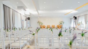 Een ruime kamer met rijen witte stoelen en een podium waarop een bloemstuk staat.