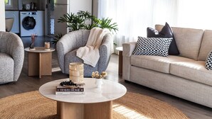 Una sala de estar moderna con un sofá gris, una mesa redonda y un sillón gris. Hay una mesita auxiliar con una vela y un libro titulado 'TRAVEL HOME'.