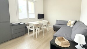 Un espace de vie moderne avec un canapé gris, une table à manger avec des chaises, un téléviseur à écran plat fixé au mur et une fenêtre avec des stores.