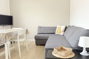 Una sala de estar con un sofá gris, una mesa blanca con mantel estampado, una cesta tejida y una lámpara blanca.