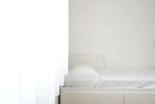 A white bed with a headboard, a white wall, and a window with sheer curtains.
