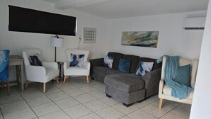 Una sala de estar con un sofá gris, dos sillones blancos, una mesita, una lámpara de pie y un televisor montado en la pared.