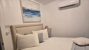 Una cama bien hecha con cabecera, una mesita de noche con control remoto y un aire acondicionado instalado en la pared.