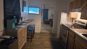 Una cocina compacta con cafetera, un televisor mostrando una escena de playa, una zona de comedor con banco y vistas al salón.