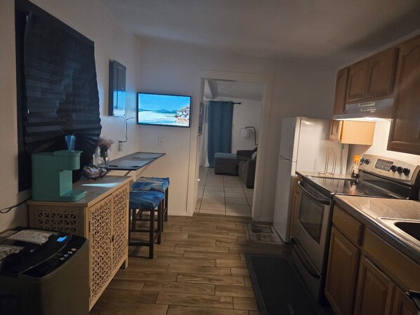 A compact kitchen with a coffee machine, a TV displaying a beach scene, a dining area with a bench, and a view of the living room.