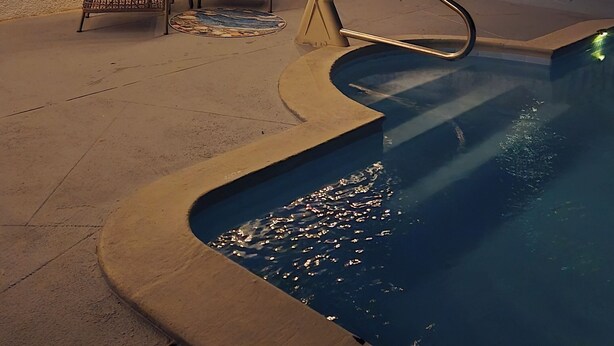 Una piscina con borde curvo, rodeada por una terraza de hormigón. Hay dos sillas de mimbre con cojines y una mesa pequeña con una botella. Luces de cuerda cuelgan sobre las sillas.