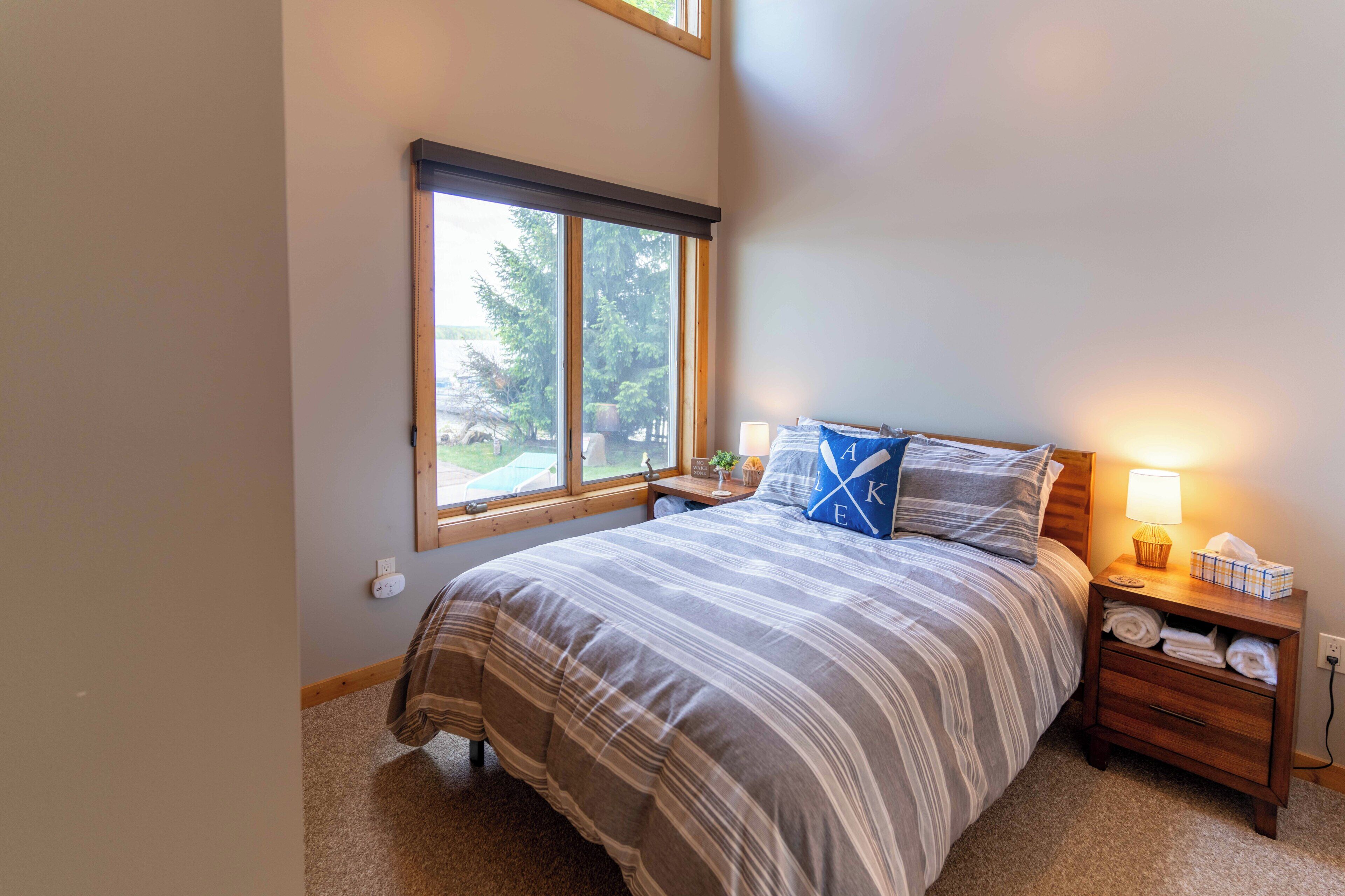 A bedroom with a large window, a bed with striped bedding, a nightstand with a lamp, and a bedside table with a tissue box.
