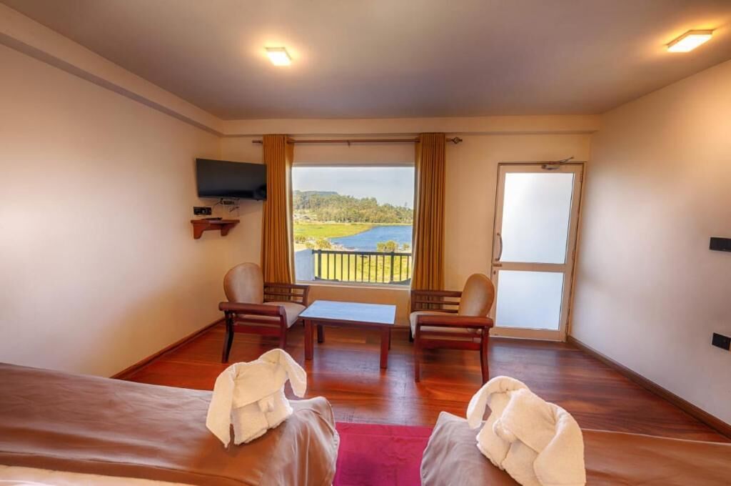 Habitación de hotel con suelo de madera, dos sillas, una mesita, una cama, un televisor montado en la pared y un balcón con vistas a un lago y colinas.