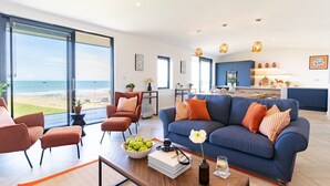 A modern living room with a large sofa, armchairs, and a coffee table. There is a view of the beach through large windows.