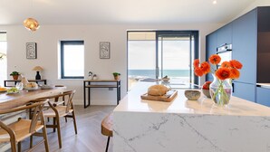 A modern kitchen with a marble countertop, wooden chairs, and a view of the sea.