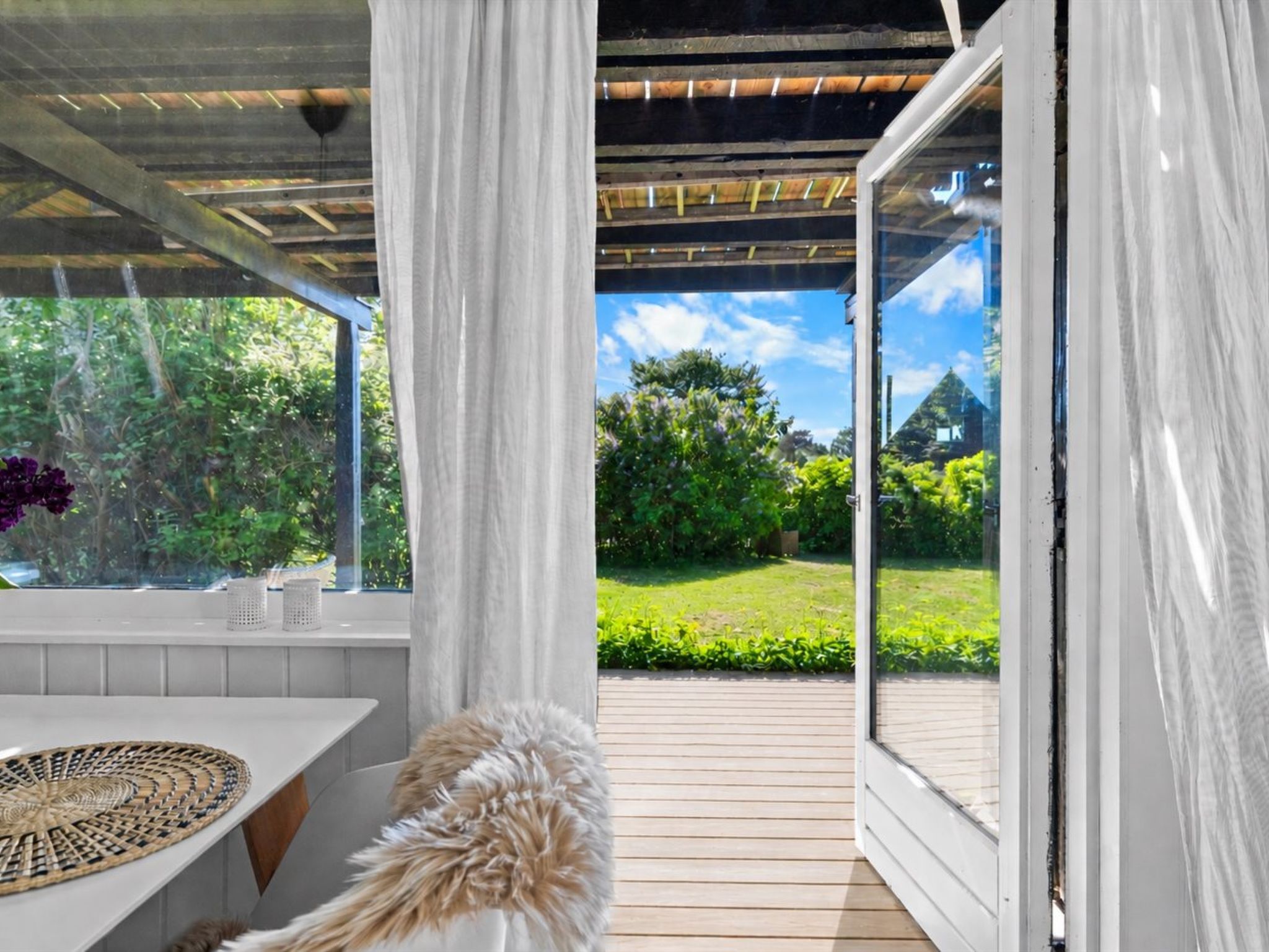 Un espacio exterior techado con cubierta de madera, una mesa blanca con centro decorativo tejido, y vista a un paisaje verde a través de un ventanal de vidrio.