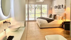 A modern bedroom with a large bed, a round mirror, a white basin, and a view of greenery through the window.