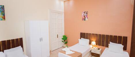 A hotel room with two beds, a white wardrobe, a nightstand with a lamp, and a decorative plant.