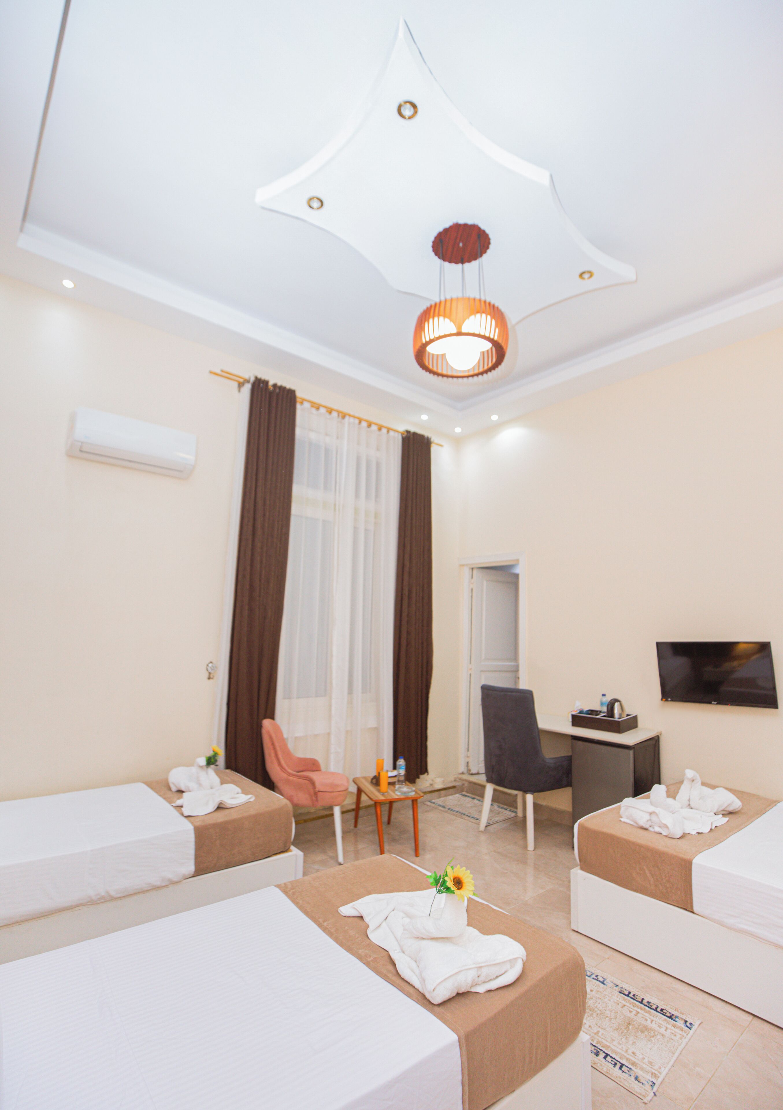 A hotel room with two beds, a desk, a chair, a TV, and a ceiling light fixture.
