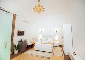 A hotel room with a bed, bedside tables, a chair, a desk, a plant, a TV, and a chandelier.