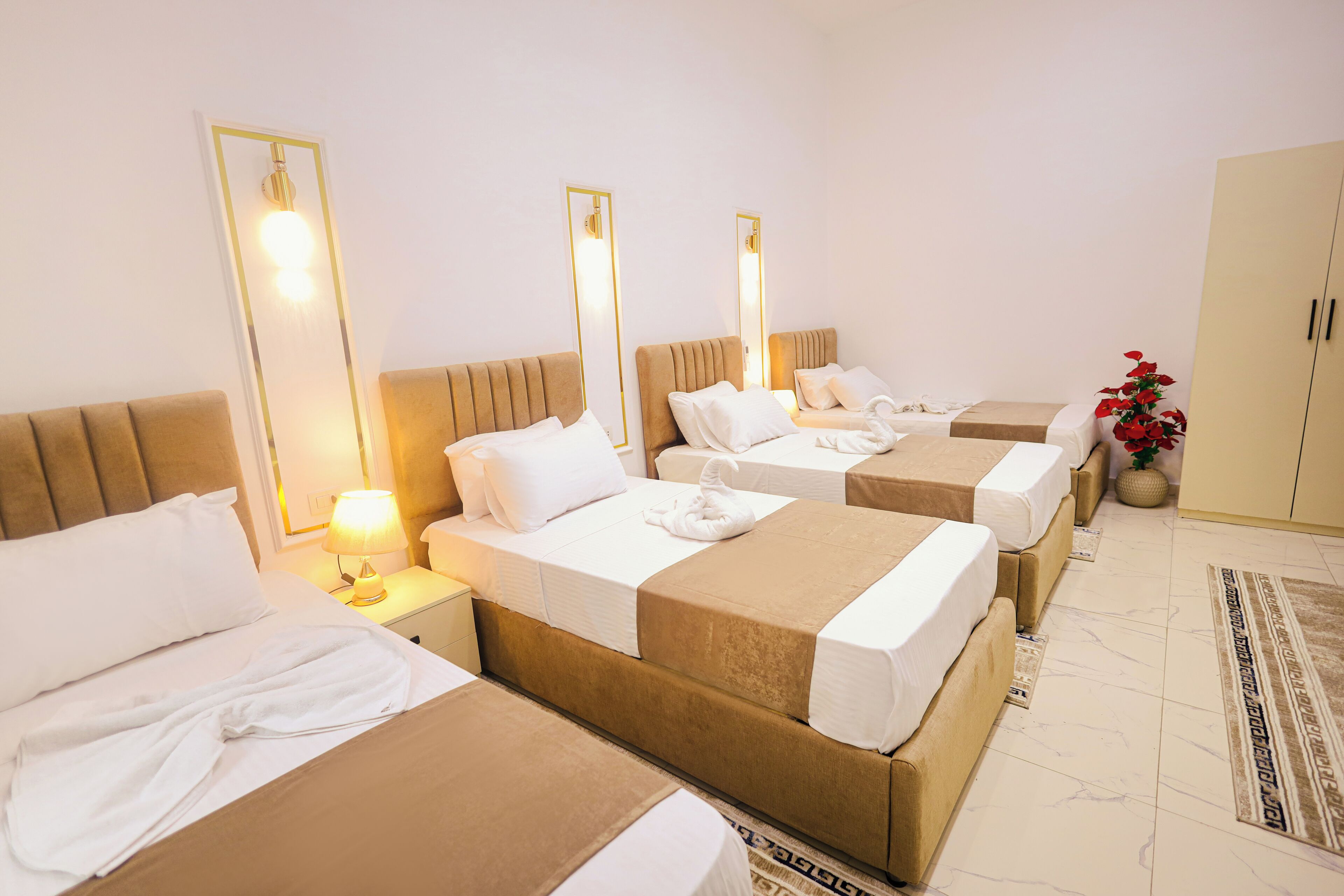 A hotel room with three beds, each with a headboard and bedside tables. There is a wardrobe and a potted plant in the corner.
