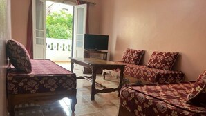 Ein Zimmer mit einer Couch, einem Bett und einem Fernseher. Ein Fenster mit Vorhängen und eine Tür, die nach draußen führt.