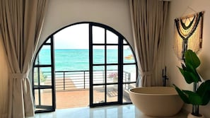 A room with a large arched window overlooking the sea, a large bathtub, and a wall-mounted macrame decoration.