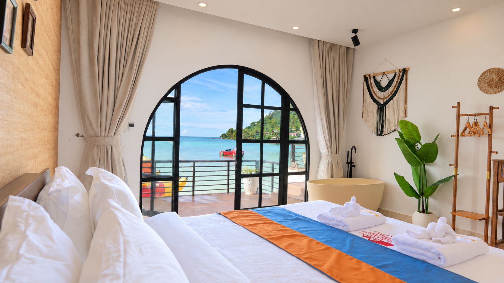 Habitación de hotel con una cama grande, vistas al mar, un balcón con vistas al mar, una planta y un perchero de madera.