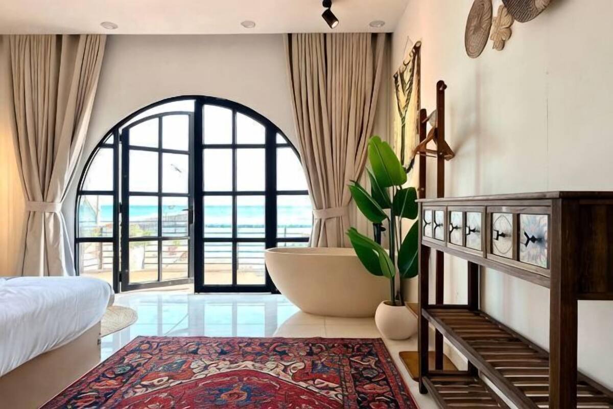 A bedroom with a large arched window, a bathtub, a bed, a wooden bench, and a potted plant.