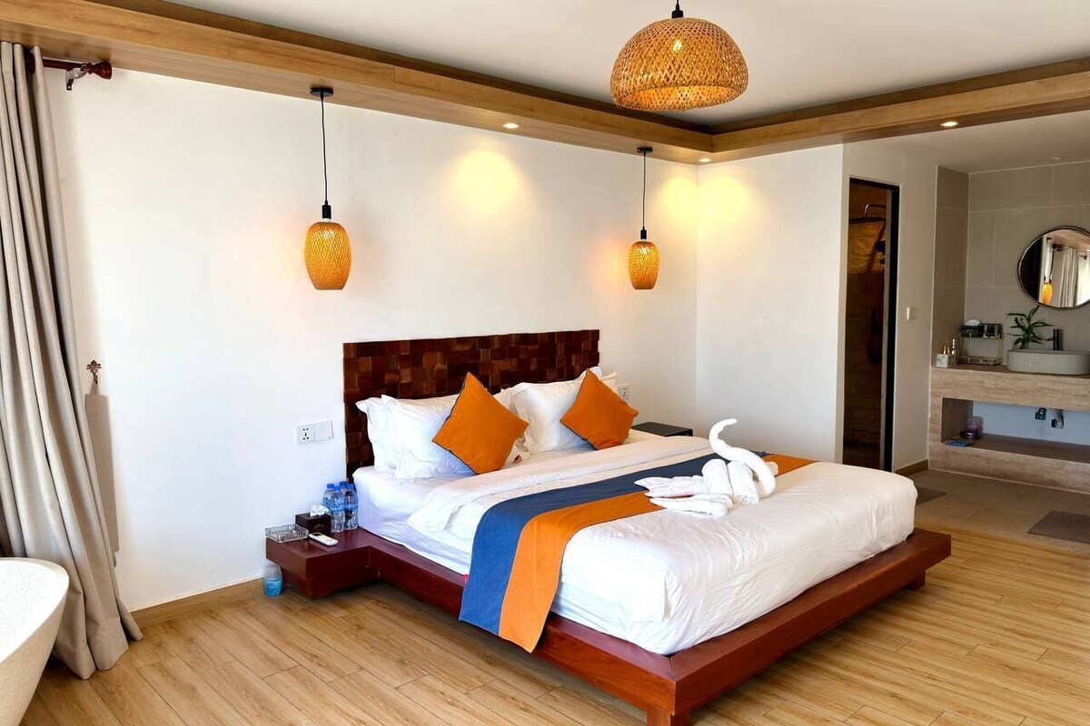 A hotel room with a large bed, wooden headboard, two bedside lamps, a bedside table, a mirror, and a bathroom with a sink and mirror.