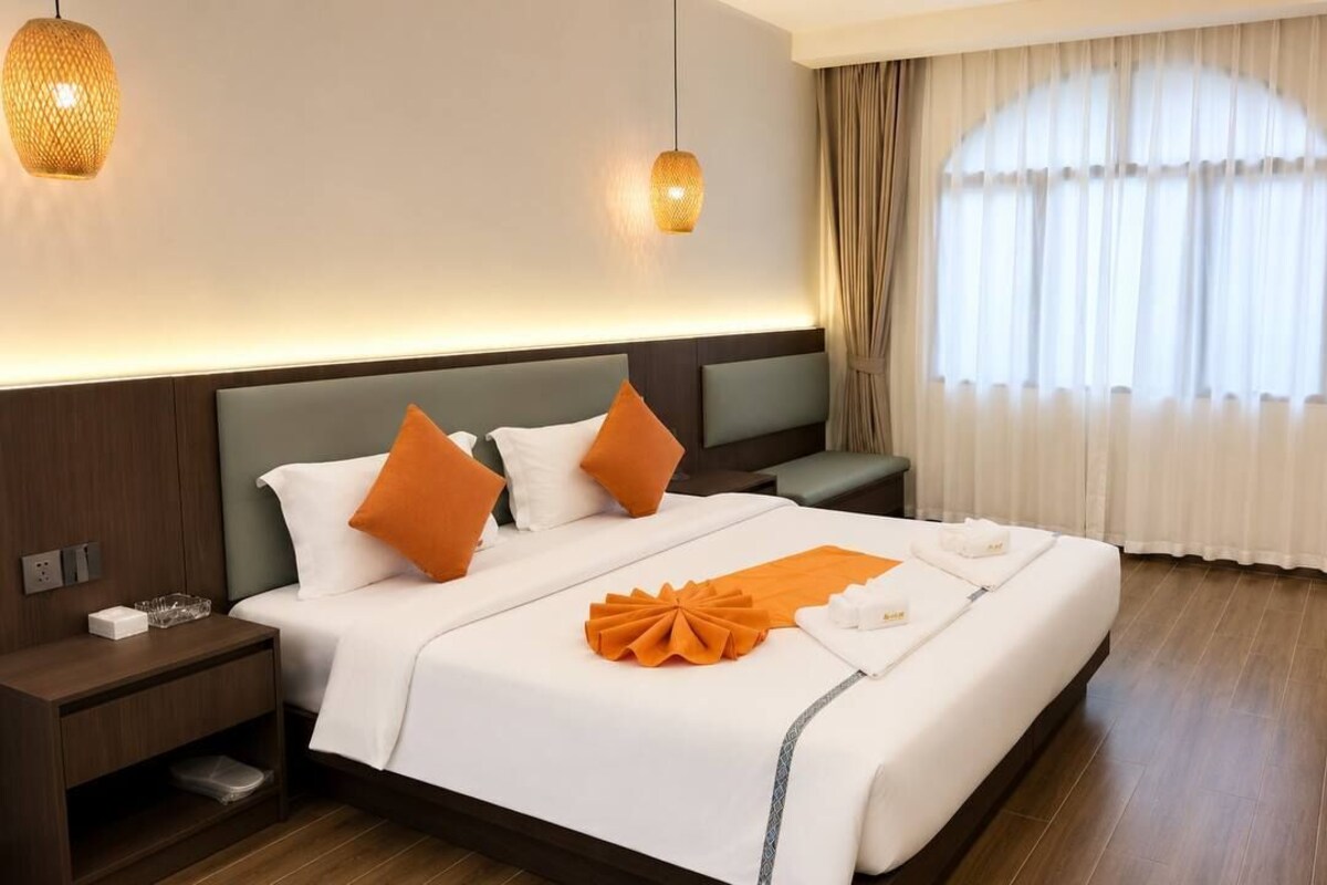 A hotel room with a large bed, bedside tables, a nightstand, a window with curtains, and two hanging lamps.