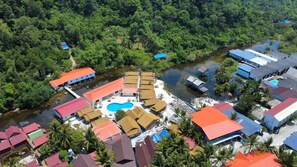 An aerial view of a resort with multiple bungalow-style accommodations, a central swimming pool, and a river running through the property.