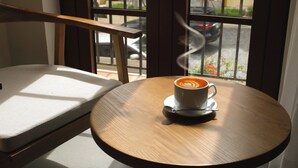 Una mesa de madera redonda con una taza blanca de café y una cuchara encima, ubicada cerca de una ventana con vista a una calle y coches estacionados.