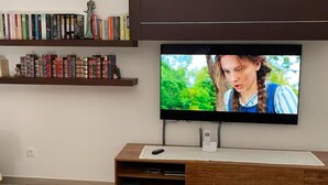 A modern living room with a flat-screen TV mounted on a dark wall above a wooden entertainment unit. Below the TV, a white remote control rests on a white paper.