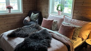 Un dormitorio de cabaña acogedor con una cama grande, dos ventanas con cortinas, una silla y una mesita.