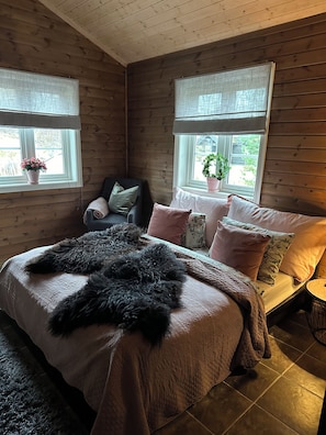 Ein gemütliches Schlafzimmer in einer Hütte mit einem großen Bett, zwei mit Vorhängen versehenen Fenstern, einem Sessel und einem kleinen Tisch.