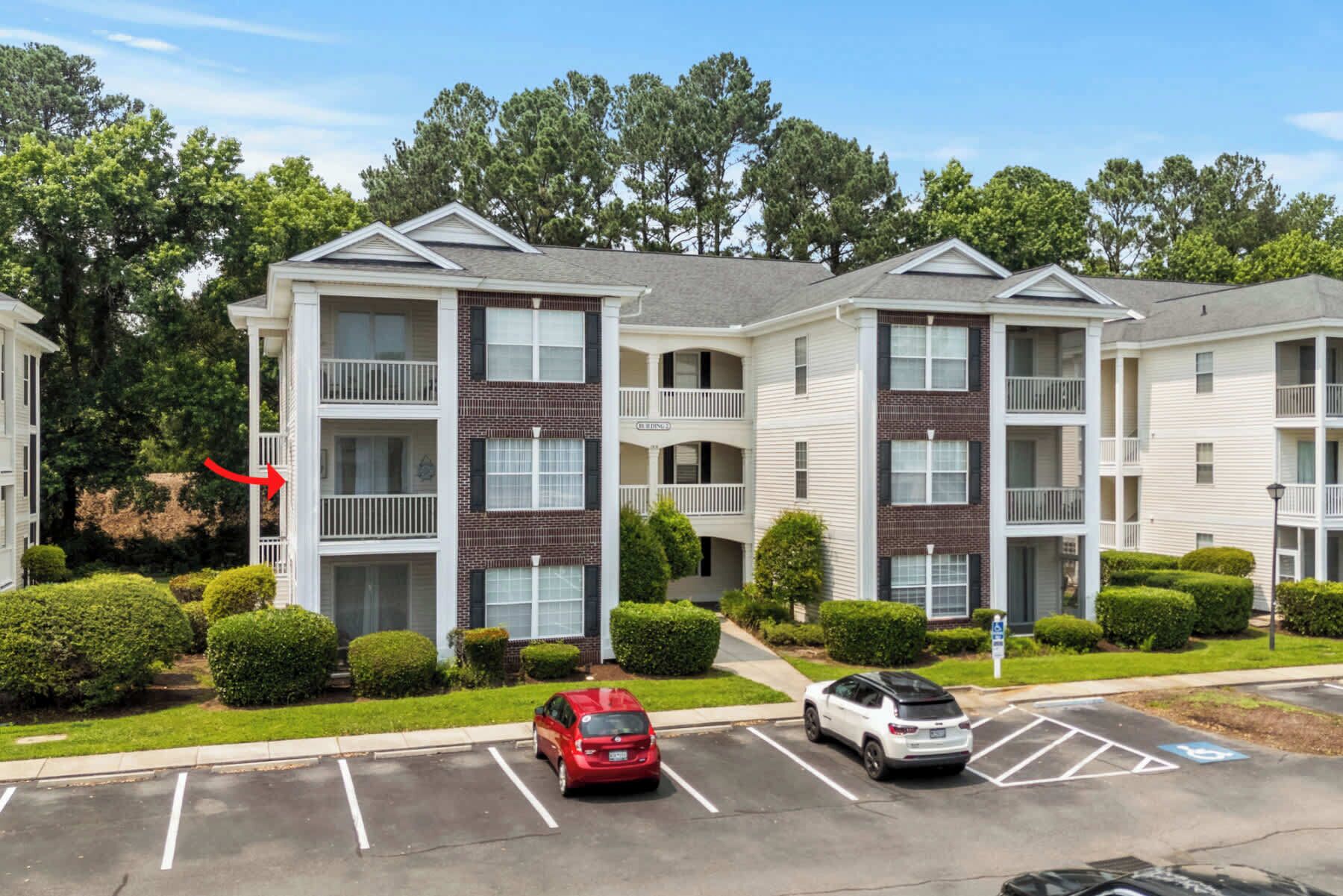 A multi-story apartment building with a brick facade, balconies, and a parking lot with two cars.