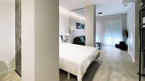 Ein modernes Hotelzimmer mit einem großen Bett, einem Schreibtisch, einem Sessel, einem Fernseher und Blick auf die Stadt durch das Fenster.