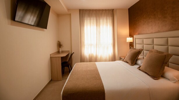 A hotel room with a bed, a desk, a chair, a TV mounted on the wall, and a window with curtains. - HOSTAL AGORA (Novetlè)