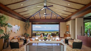 A spacious indoor area with a view of the ocean, featuring a central seating area with sofas and a coffee table, surrounded by potted plants and artwork on the walls. - Laueki 5 – Ocean View Estate | Onsite Staff & Daily Housekeeping (Kailua Kona)