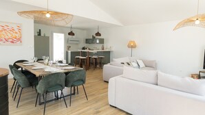 Eine moderne Küche mit weißen Schränken und einer zentralen Kochinsel, ein Essbereich mit Holztisch und -stühlen sowie ein Wohnbereich mit weißem Sofa und grünen Sesseln.