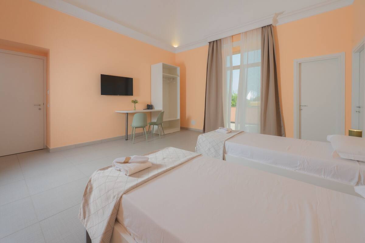A hotel room with two beds, a desk, a TV, and a window with curtains.