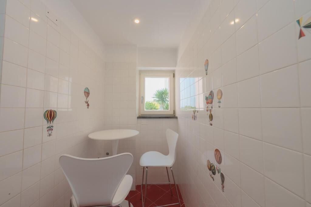 Une petite salle de bain lumineuse, avec des carreaux blancs, une fenêtre, une chaise blanche et une petite table.