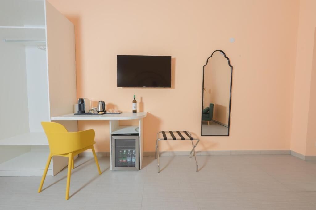 A small, modern room with a white desk, a yellow chair, a mini-fridge, a television, and a mirror.