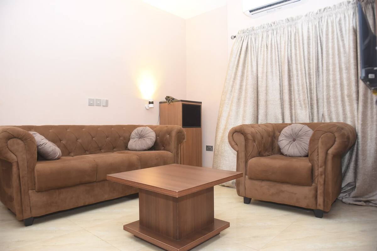 Un salon avec deux canapés marron, une table basse en bois, un téléviseur fixé au mur et un rideau.