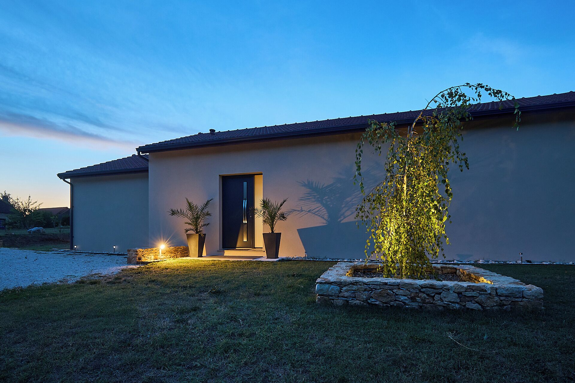 Una casa moderna con una puerta oscura, dos plantas en macetas y una jardinera de piedra con un árbol.