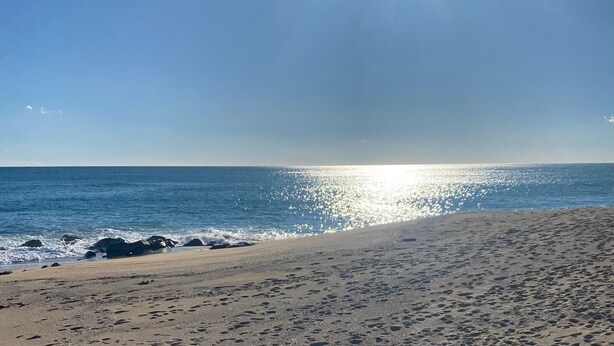 A beach with footprints leading to the water, a clear sky, and the sun shining on the horizon. - Beach apartment, in front of the sea! (Caldes d'Estrac)