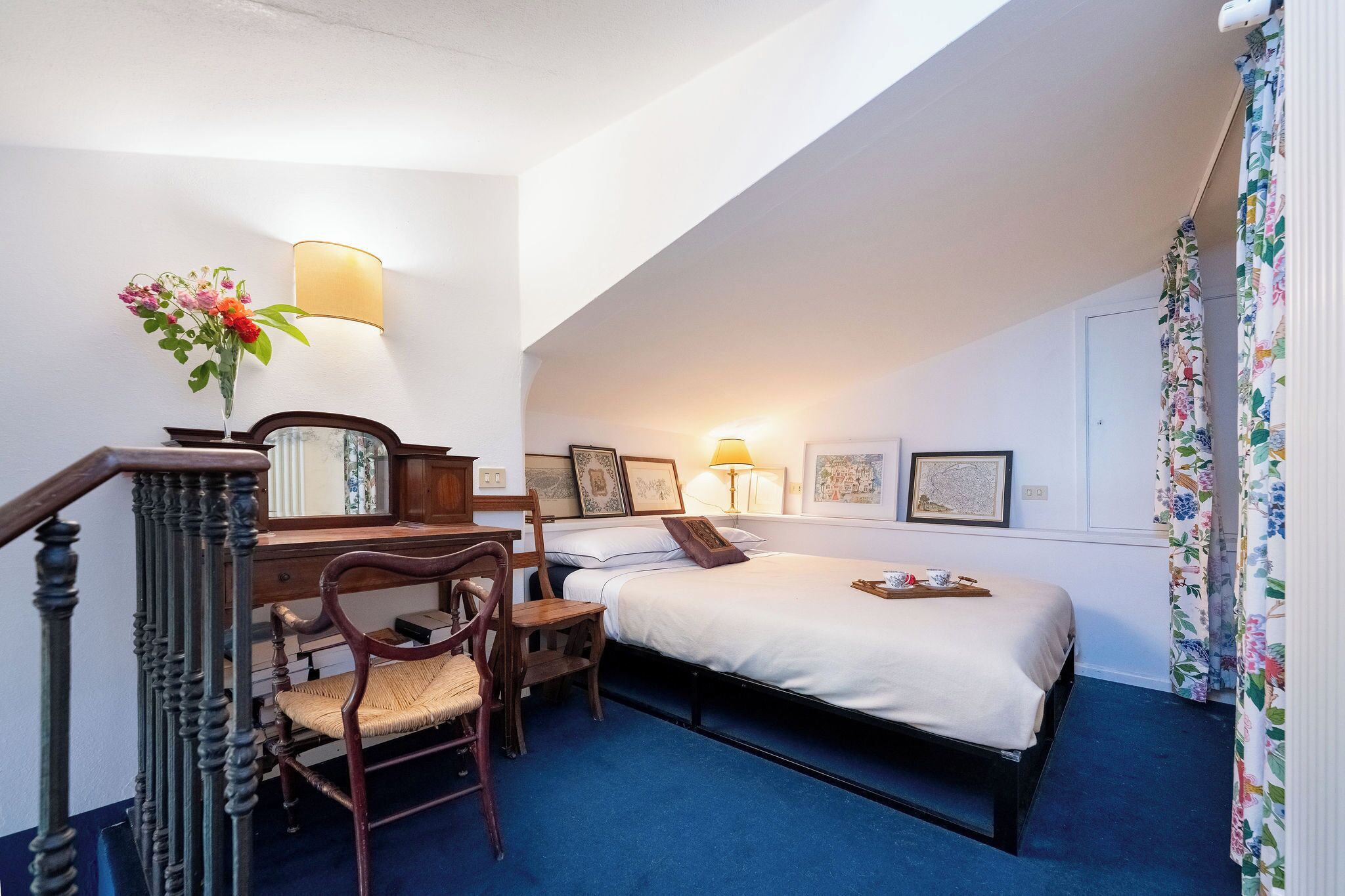 A bedroom with a bed, two chairs, a desk, a lamp, and a vase with flowers.